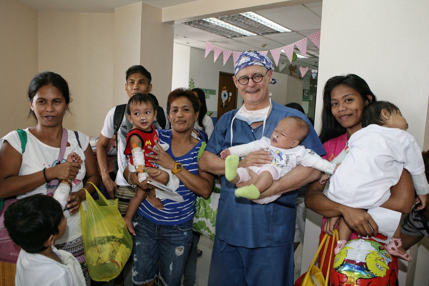 A volunteer surgeon in theatre scrubs holds a baby, surrounded by smiling families