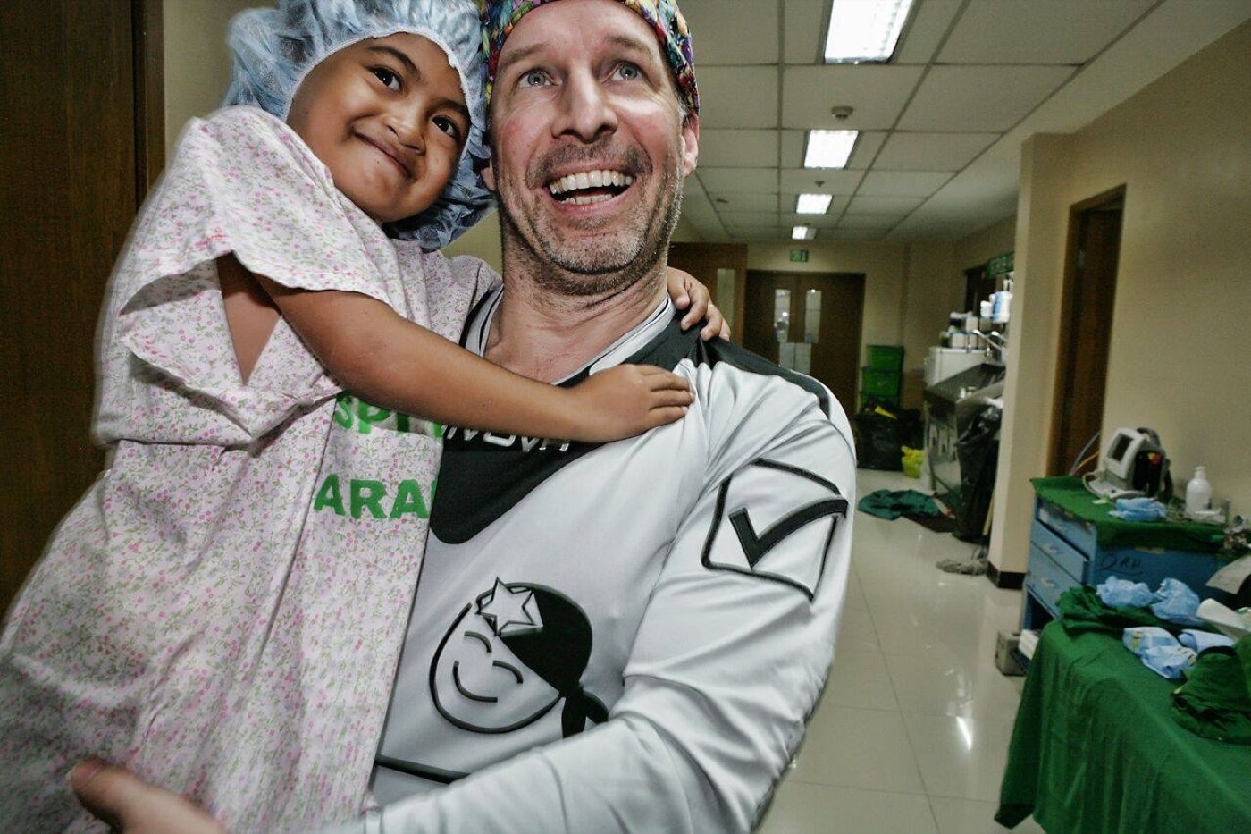 A young girl in a surgical cap smiles in the arms of a volunteer after her operation